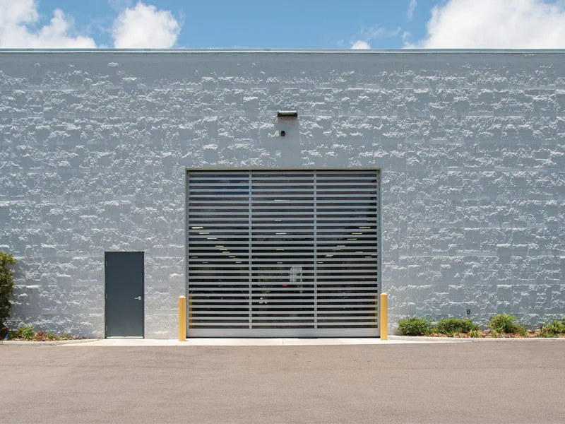 Commercial garage door with horizontal slats, gray exterior wall, and adjacent pedestrian door, illustrating design versatility and functionality for commercial applications.