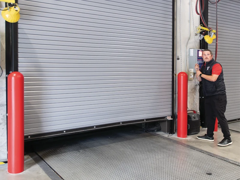 Commercial technician operating a motorized overhead garage door in an industrial setting, featuring safety bollards and control panel, emphasizing professional garage door replacement services by Overhead Door Company of The Desert™.