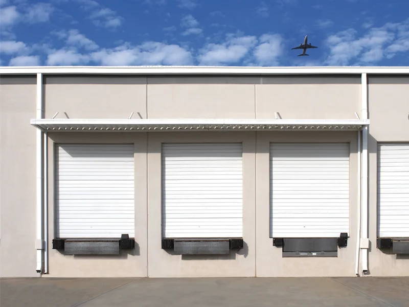 Commercial garage doors with white rolling shutters on a warehouse, featuring loading docks and a blue sky with clouds above.