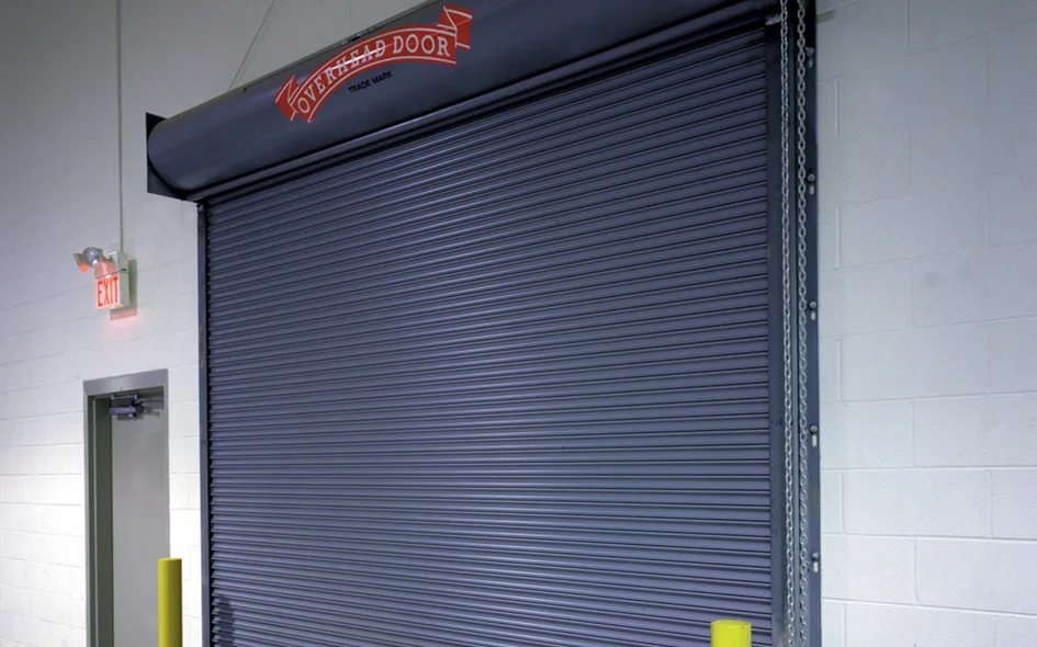 Fire-rated rolling service door model 631 by Overhead Door Company, featuring a sleek gray curtain and safety yellow bollards, installed in a commercial setting.